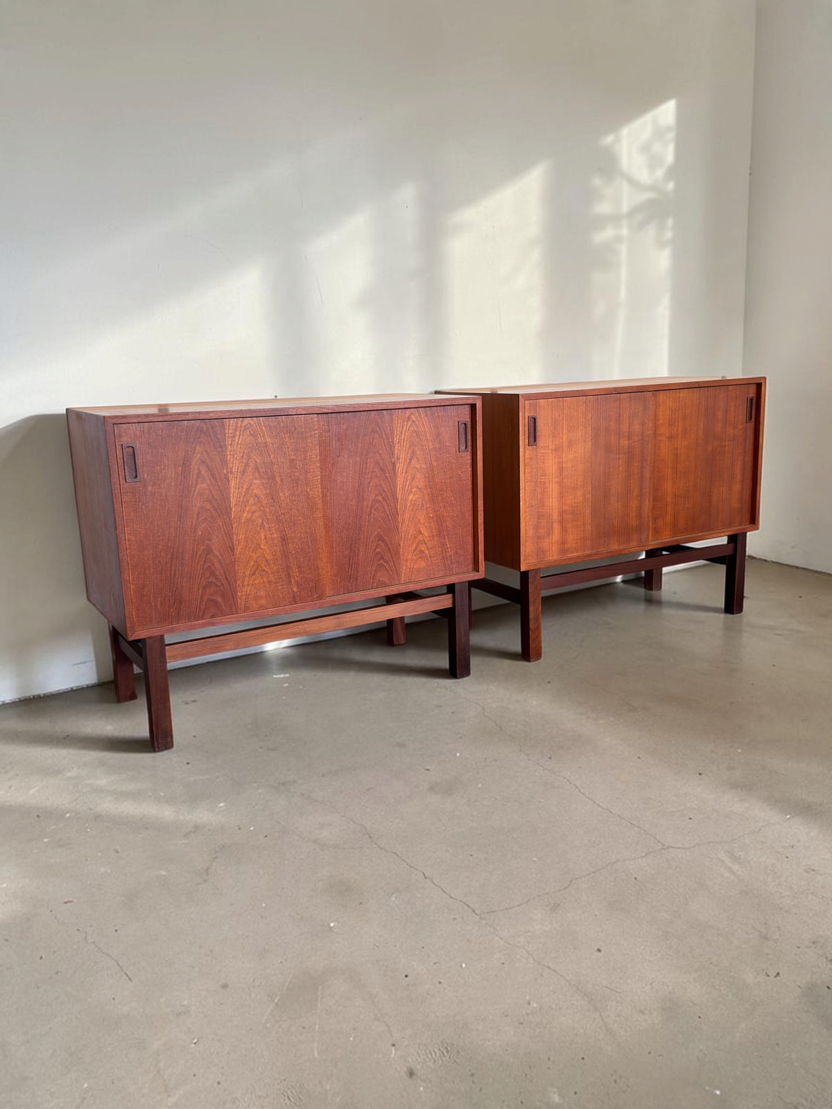 Twee tessvintage Deense teak dressoirs met schuifdeuren en een donker houten H-frame onderstel in een lichte ruimte.