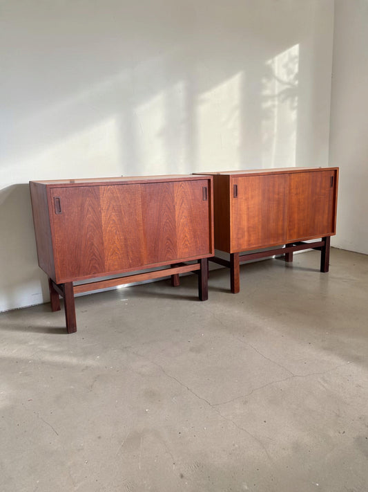 Twee tessvintage Deense teak dressoirs met schuifdeuren en een donker houten H-frame onderstel in een lichte ruimte.